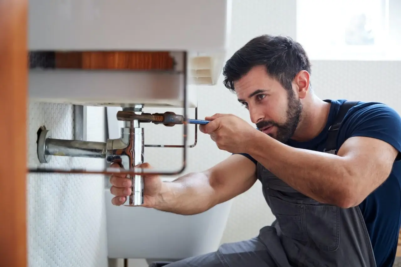 Plumber in a dark blue shirt tightening pipes under a sink with a wrench.