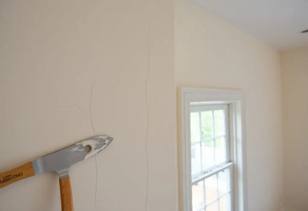 Beige interior wall with vertical cracks and a hammer resting against it, suggesting renovation in progress near a window.