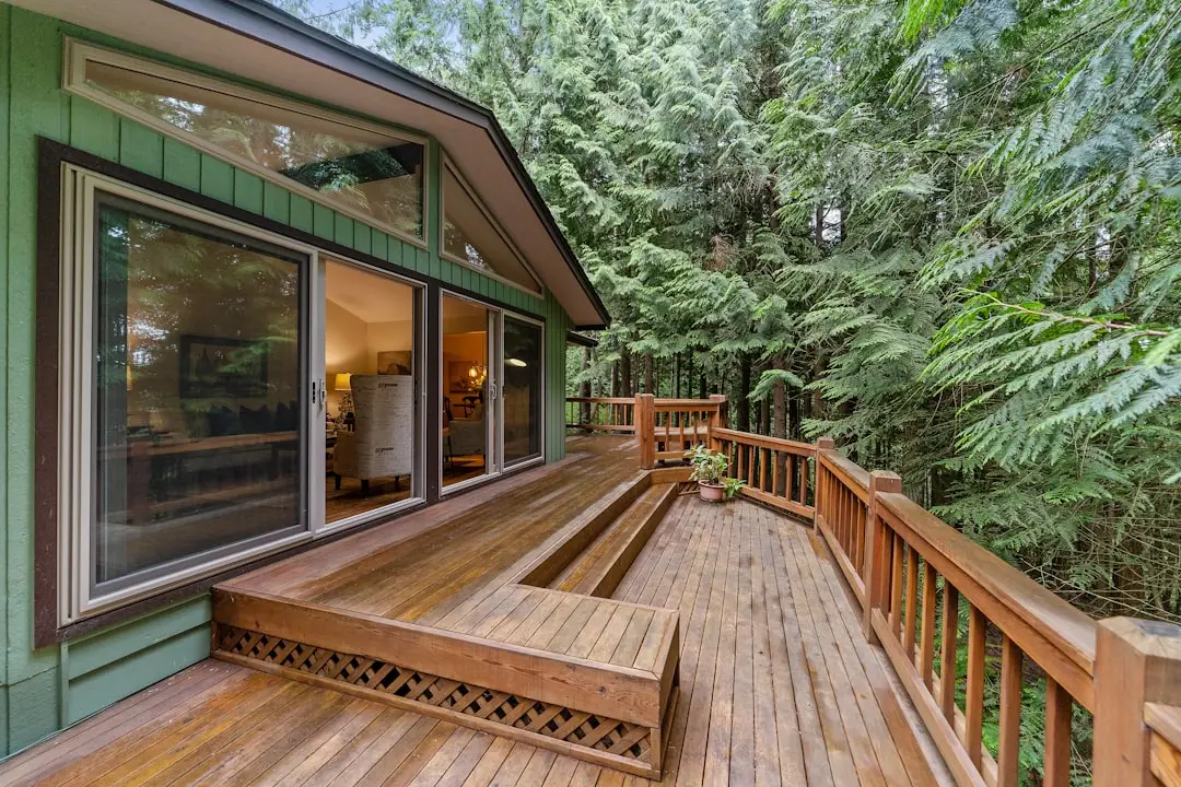 Green cabin with large sliding glass doors opening to a warmly lit living room, adjacent to a wooden deck in a forest setting.