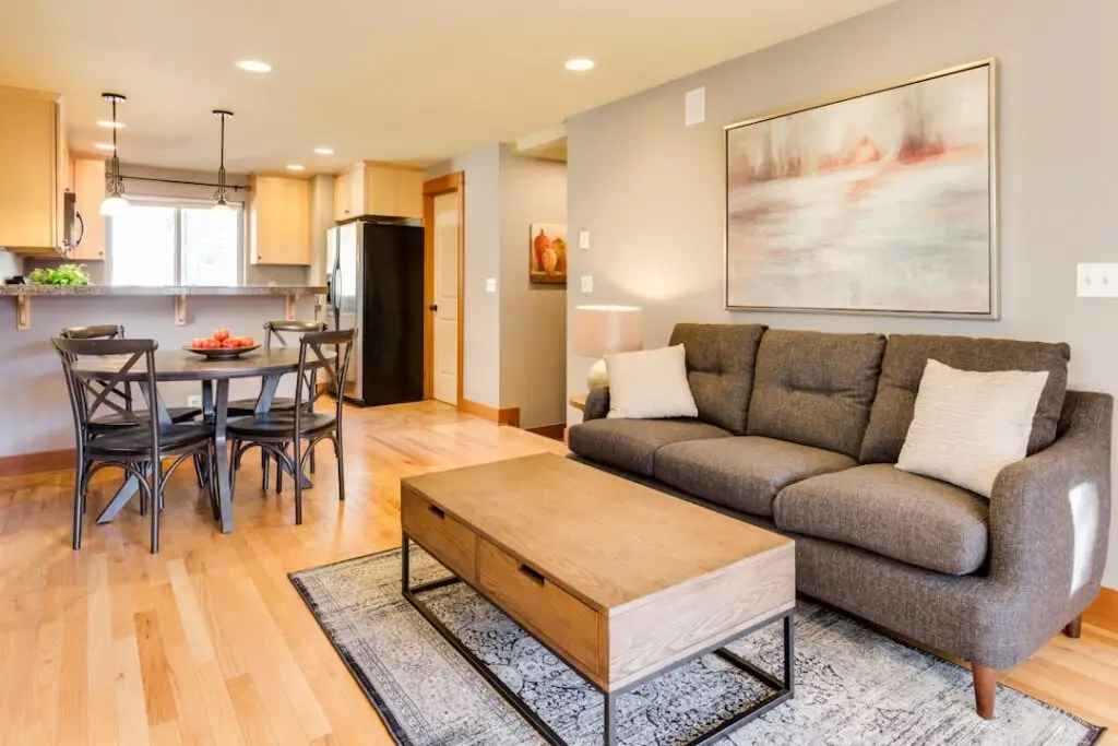 Gray sofa with white pillows, wooden coffee table, and rug in an open living area beside a dining table and kitchen.