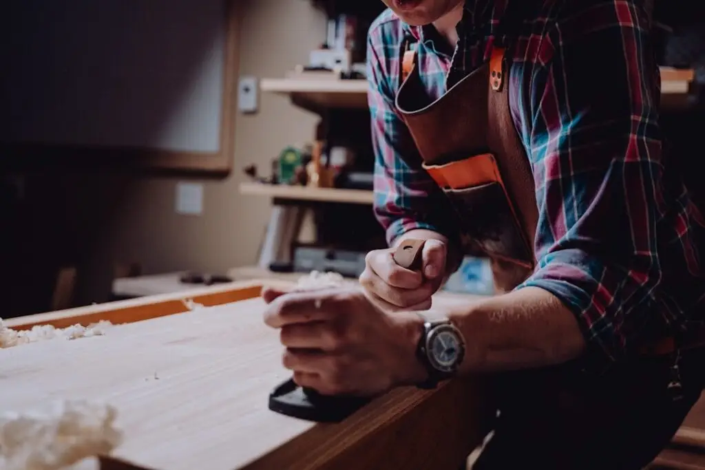 Woodworker in a plaid shirt and apron planes a wooden plank on a workbench, concentrating on shaping the edge.